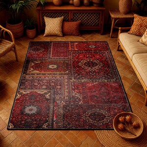 May include: A large, rectangular area rug with a patchwork design. The rug features a deep red and burgundy color palette with intricate, traditional patterns. The rug is placed on a terracotta tile floor, with a wicker chair and sofa in the background.