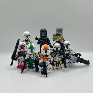 May include: A group of Lego clone trooper minifigures, each with distinct helmet and armour designs. The figures are holding black blasters, and the colour variations include white, black, orange, green, and yellow. The figures are on a plain background.