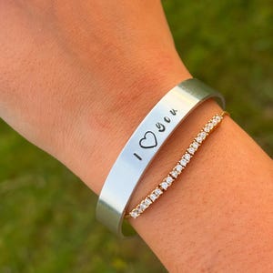 May include: A silver cuff bracelet engraved with "I ❤️ you", alongside a gold-coloured tennis bracelet. The bracelets are displayed on a wrist, set against a blurred green backdrop.