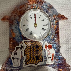 May include: A decorative clock with a white face and black Roman numerals. The clock is encased in a sparkly resin with blue, copper, and gold tones. Baseball team stickers are on the clock.
