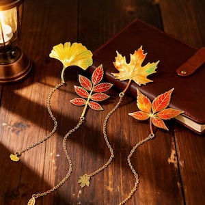 Può includere: Quattro segnalibri a forma di foglia in metallo nei toni del giallo, arancione e rosso con catene dorate. I segnalibri sono su una superficie di legno accanto a un taccuino rilegato in pelle marrone e una lanterna accesa.