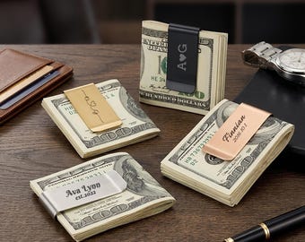 Puede incluir: Varias pilas de billetes de cien dólares estadounidenses se sujetan con pinzas para billetes personalizadas. Las pinzas son de varios colores, como negro, dorado y plateado, y tienen nombres y fechas grabados. También se ve una cartera de cuero marrón y un reloj de pulsera.