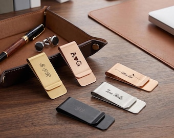 Puede incluir: Varios clips de dinero personalizados de metal en dorado, oro rosa, plateado y negro. Cada clip presenta iniciales o nombres grabados, incluyendo "A♥G", "E&R", "Mia Lee", "Zoe Belle" y "J.L.".