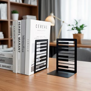 Puede incluir: Soportes para libros de metal negro con diseño de teclas de piano, sosteniendo una pila de libros. Los libros tienen cubiertas blancas con títulos como "CEREAL" e "ISABEL MARANT". Los sujetalibros están sobre una superficie de madera.