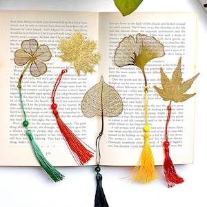 May include: Gold-colored metal leaf-shaped bookmarks with colorful tassels. The bookmarks are in various leaf designs, including a maple leaf, ginkgo leaf, and a four-leaf clover, resting on the pages of an open book.