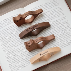 Può includere: Quattro fermapagina in legno con i nomi Ursulina, Seraphina, Theodore e Rosalind. Ognuno ha un design unico, tra cui una faccia di gatto e dettagli floreali. Sono appoggiati su un libro aperto.