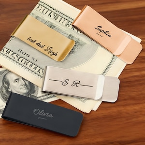 Puede incluir: Una variedad de pinzas para billetes personalizadas en oro, oro rosa, plata y negro. Las pinzas están grabadas con nombres y frases como "best dad Leigh" e iniciales. Las pinzas para billetes se muestran sobre billetes estadounidenses.
