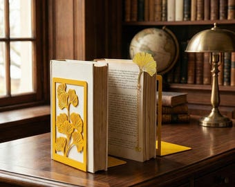 Könnte beinhalten: Ein Paar gelbe Metall-Buchstützen mit Ginkgo-Blatt-Design, die ein Buch halten. Ein passender Ginkgo-Blatt-Lesezeichen befindet sich im Buch. Die Buchstützen und das Lesezeichen stehen auf einem Holzschreibtisch.