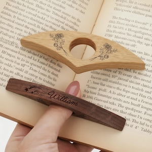 Puede incluir: Dos accesorios de libro de madera: un sujetap&aacute;ginas de roble claro con grabado floral y un marcap&aacute;ginas de nogal oscuro con el nombre "William" y detalles de estrellas. Ambos est&aacute;n sobre un libro abierto.