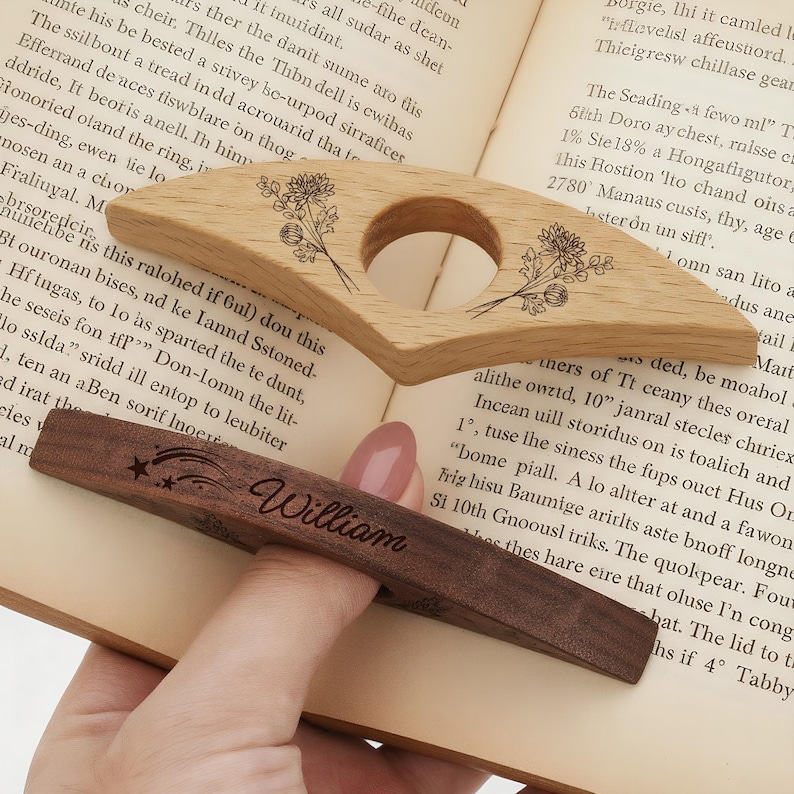 Puede incluir: Dos accesorios de libro de madera: un sujetap&aacute;ginas de roble claro con grabado floral y un marcap&aacute;ginas de nogal oscuro con el nombre "William" y detalles de estrellas. Ambos est&aacute;n sobre un libro abierto.