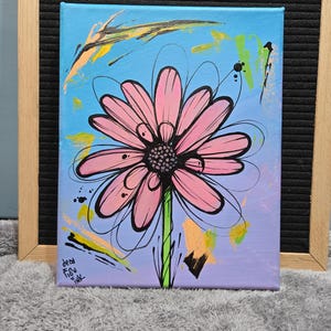 Puede incluir: Pintura sobre lienzo de una gran margarita rosa con contorno y centro negros. El fondo es un degradado de azul y morado con salpicaduras de pintura de colores. La pintura está en un marco de madera.