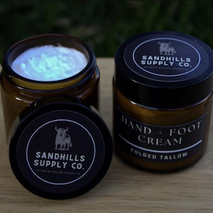 May include: Three amber glass jars of Sandhills Supply Co. hand and foot cream. One jar is open, revealing a white cream. The labels are black with white text and a bull logo. The text reads "Hand + Foot Cream" and "Folded Tallow".