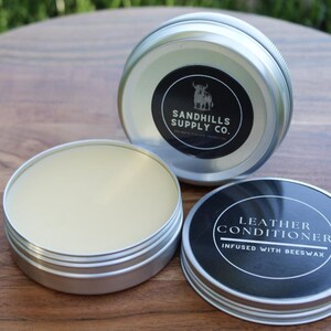 May include: Three open silver tins of leather conditioner on a wooden surface. One tin is open, revealing a light yellow balm. The lids feature black labels with white text and a bull logo. One label reads "Leather Conditioner infused with beeswax."