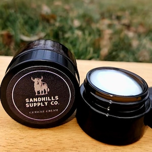 May include: Two black jars of cuticle cream. One jar is closed, displaying a label with a bull illustration and the text "SANDHILLS SUPPLY CO. CUTICLE CREAM." The other jar is open, revealing a white cream. The background is blurred.