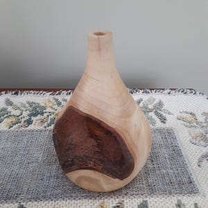May include: A wooden vase with a teardrop shape. The vase is made of light-coloured wood with a large, dark brown patch on the side. The vase has a smooth, polished finish and a small opening at the top.