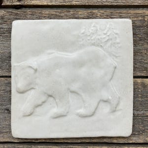 May include: A square, off-white ceramic tile featuring a relief of a bear walking to the left. Trees and a sun are in the upper right corner. The tile is set against a weathered wood background.