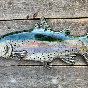 May include: A ceramic wall sculpture shaped like a trout, featuring a glossy glaze with a gradient of deep green along the back, a pinkish-purple lateral stripe, and a cream-colored underbelly covered in small, dark, distinct black spots.