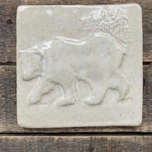 May include: A square, off-white ceramic tile featuring a raised relief of a bear walking to the left. The bear is depicted with detailed fur and a tree is behind the bear. The tile has a speckled texture and rounded edges.