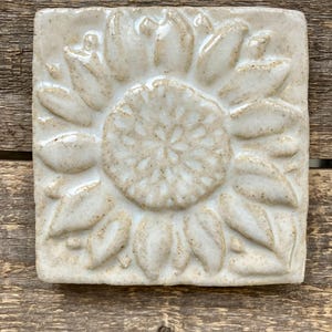 May include: A square ceramic tile featuring a raised, embossed sunflower design. The tile has a glossy, off-white glaze with visible speckles throughout the surface, highlighting the textured petals and the detailed center of the flower motif.