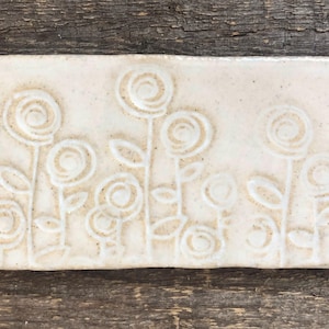 May include: A rectangular, off-white ceramic tile with an embossed floral design. The design features stylized flowers with spiral centers and slender stems, and is set against a textured background. The tile is displayed on a weathered wooden surface.