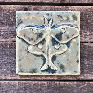 May include: Square ceramic tile with a raised design of a moth. The tile has a mottled, textured surface with shades of green, blue, and white. The moth design is in a darker blue-green color, with detailed wing and body features.