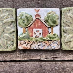 May include: Three square ceramic tiles. Two tiles feature a sunflower design in shades of green and cream. The center tile depicts a red barn with green trees and a white sky. The tiles are set on a weathered wooden surface.