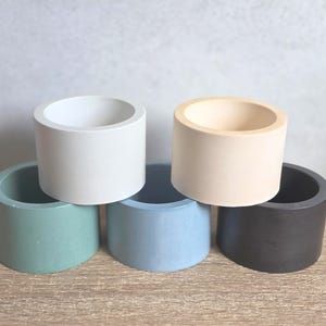 May include: Six small, cylindrical concrete planters in a range of colours: white, beige, green, blue, and dark grey. The planters are displayed on a wooden surface, highlighting their simple, modern design.