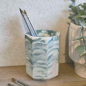 May include: A hexagonal pen holder with a marbled blue and white design. Several clear pens with brown ink are inside. The holder is on a wooden surface, with a plant in a white pot in the background.