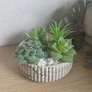 May include: A small, round, ribbed planter with a cream and black marbled finish, filled with white decorative stones and an arrangement of various artificial green succulents in shades of light and dark green with subtle purple tips.