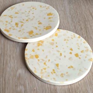 May include: Two circular coasters featuring a white base embedded with scattered yellow and translucent fragments. The coasters have a smooth, flat surface and a clean, rounded edge, showcasing a speckled terrazzo-style pattern throughout.
