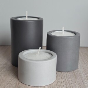 May include: Three cylindrical candle holders in varying heights and shades of grey. The tallest is dark charcoal, the medium is mid-tone grey, and the shortest is light grey. Each holder contains a white wax candle with a centred cotton wick.