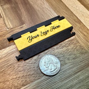 May include: A black and yellow cable protector with the text "Your Logo Here" is displayed on a wooden surface. A US quarter coin is next to the cable protector.