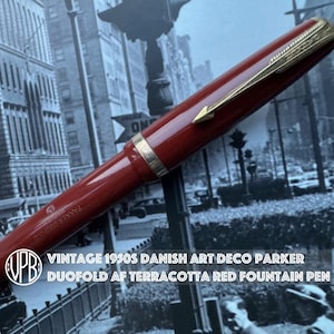 Könnte beinhalten: Vintage dänischer Art Deco Parker Duofold AF Füllfederhalter in Terracotta-Rot aus den 1950er Jahren. Der Stift hat einen roten Korpus mit goldfarbenen Akzenten. Das Design des Stifts ist klassisch und elegant, mit zeitlosem Reiz. Der Stift ist ein Schreibgerät.