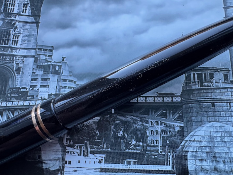 May include: Close-up of a black fountain pen with gold accents. The pen has the text "U.S. PARKER DUOFOLD REG. T. M." engraved on the barrel. The pen is set against a backdrop of a cityscape.