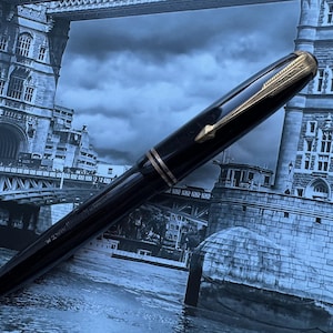 May include: A black fountain pen with gold-colored accents. The pen features a gold-colored arrow clip and a gold band near the nib. The pen is set against a backdrop of a blue-toned image of a bridge.