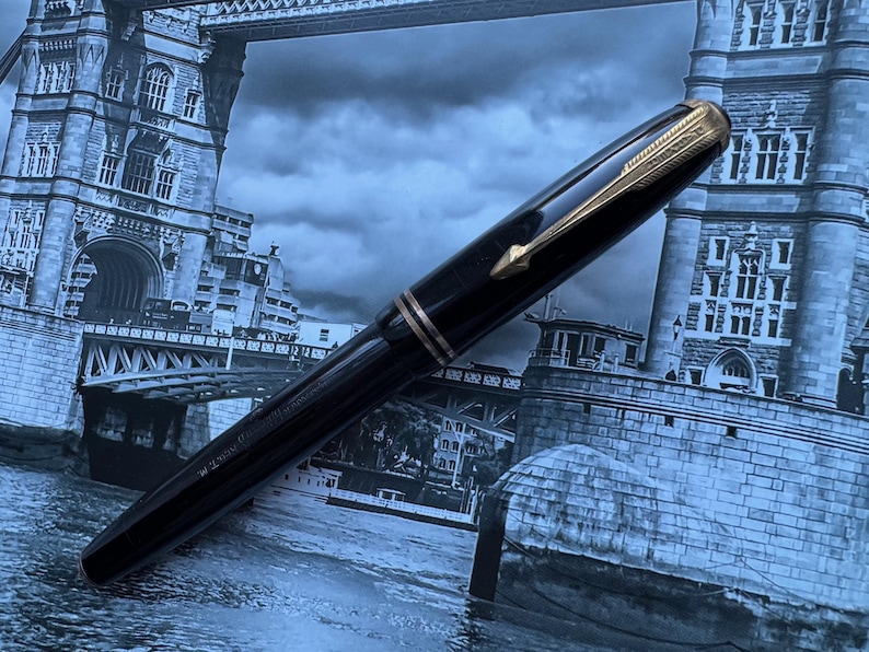 May include: A black fountain pen with gold-colored accents. The pen features a gold-colored arrow clip and a gold band near the nib. The pen is set against a backdrop of a blue-toned image of a bridge.
