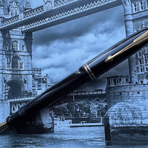 May include: A black fountain pen with gold accents, set against a backdrop of the Tower Bridge in London. The pen features a gold nib and a sleek, polished design. A classic writing instrument.