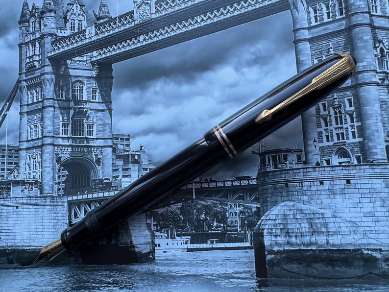 May include: A black fountain pen with gold accents, set against a backdrop of the Tower Bridge in London. The pen features a gold nib and a sleek, polished design. A classic writing instrument.