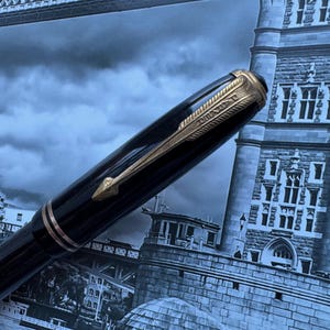 May include: A black fountain pen with gold-colored accents, including a clip and decorative bands. The pen is set against a backdrop of a London cityscape, with the Tower Bridge visible in the background.