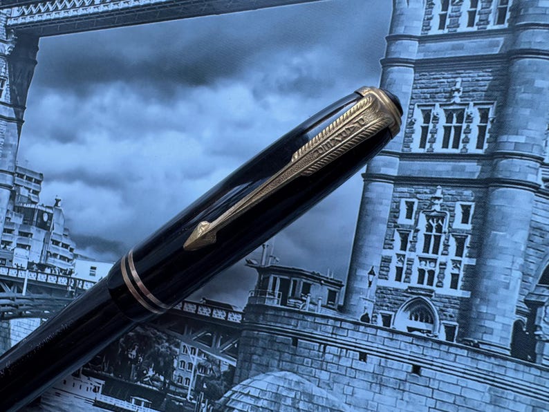May include: A black fountain pen with gold-colored accents, including a clip and decorative bands. The pen is set against a backdrop of a London cityscape, with the Tower Bridge visible in the background.
