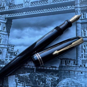 May include: A black fountain pen with gold accents, including the nib and decorative bands, lies open on a blue-toned background. The pen's cap is placed beside it, revealing the gold clip. The background features a blurred image of a bridge.