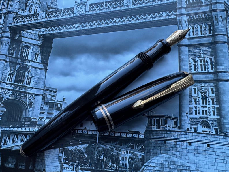 May include: A black fountain pen with gold accents, including the nib and decorative bands, lies open on a blue-toned background. The pen's cap is placed beside it, revealing the gold clip. The background features a blurred image of a bridge.