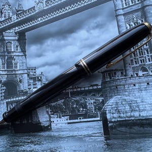 May include: A black fountain pen with gold-colored accents and a clip. The pen is set against a backdrop of the Tower Bridge in London. The pen's design includes a gold band near the nib and at the top.