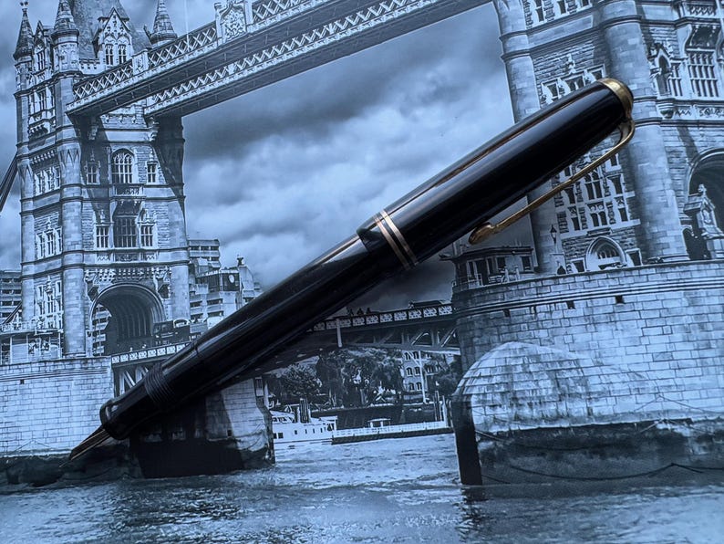 May include: A black fountain pen with gold-colored accents and a clip. The pen is set against a backdrop of the Tower Bridge in London. The pen's design includes a gold band near the nib and at the top.