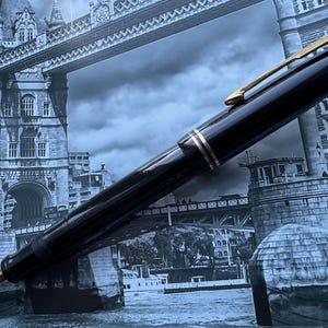 May include: A black fountain pen with gold-colored accents, including the clip and bands, is displayed against a backdrop of the Tower Bridge in London. The pen's nib is visible, suggesting it's ready for use. The pen is a classic writing instrument.
