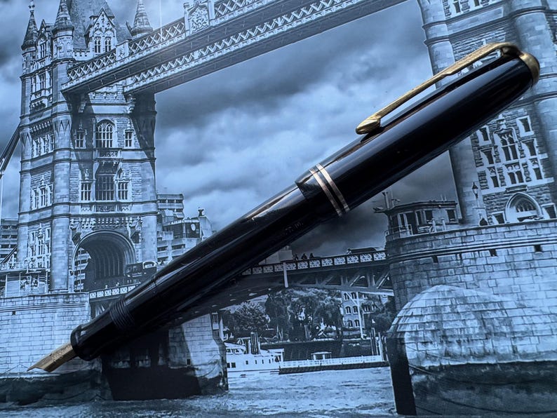 May include: A black fountain pen with gold-colored accents, including the clip and bands, is displayed against a backdrop of the Tower Bridge in London. The pen's nib is visible, suggesting it's ready for use. The pen is a classic writing instrument.