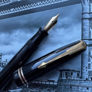 May include: A black fountain pen with a gold nib and accents. The pen's cap is off, revealing the nib and ink reservoir. The pen is set against a backdrop of a blue and gray image of a bridge.