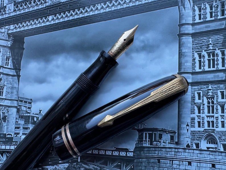 May include: A black fountain pen with a gold nib and accents. The pen's cap is off, revealing the nib and ink reservoir. The pen is set against a backdrop of a blue and gray image of a bridge.