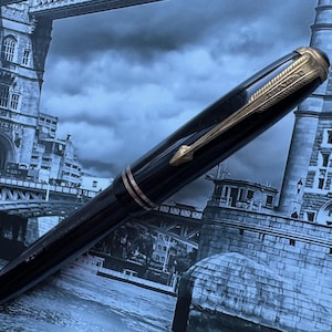 May include: A black fountain pen with gold-colored accents, including the clip and trim. The pen is set against a backdrop of a grayscale image of a bridge and buildings.