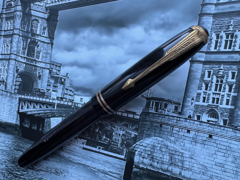May include: A black fountain pen with gold-colored accents, including the clip and trim. The pen is set against a backdrop of a grayscale image of a bridge and buildings.